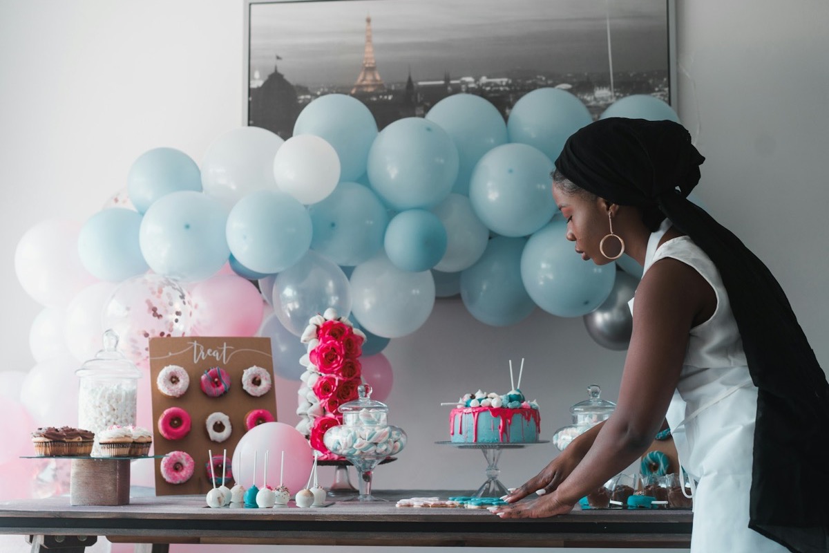 A host arranging a dessert table under a pastel balloon garland